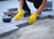 Buttrose Landscaping - person in yellow gloves lays paving stones in layers. Garden brick pathway paving by professional paver worker. Laying gray concrete paving slabs in house courtyard on sand foundation base.