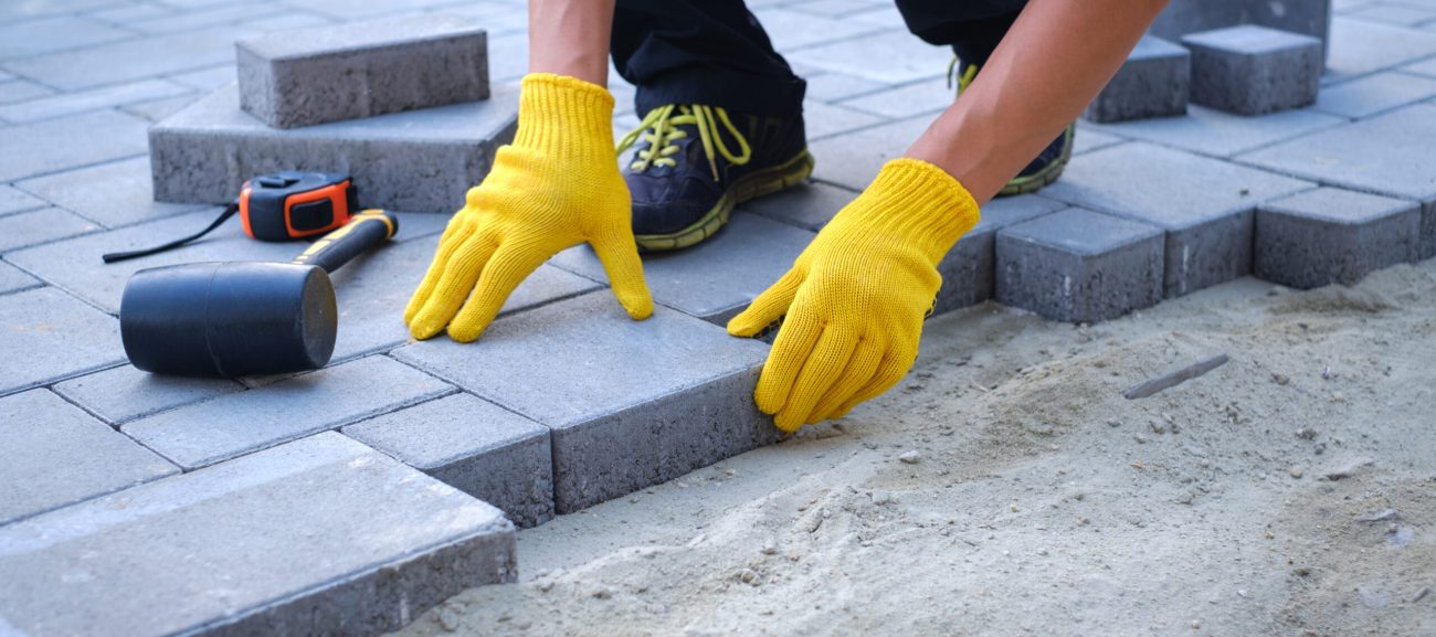 Buttrose Landscaping - person in yellow gloves lays paving stones in layers. Garden brick pathway paving by professional paver worker. Laying gray concrete paving slabs in house courtyard on sand foundation base.