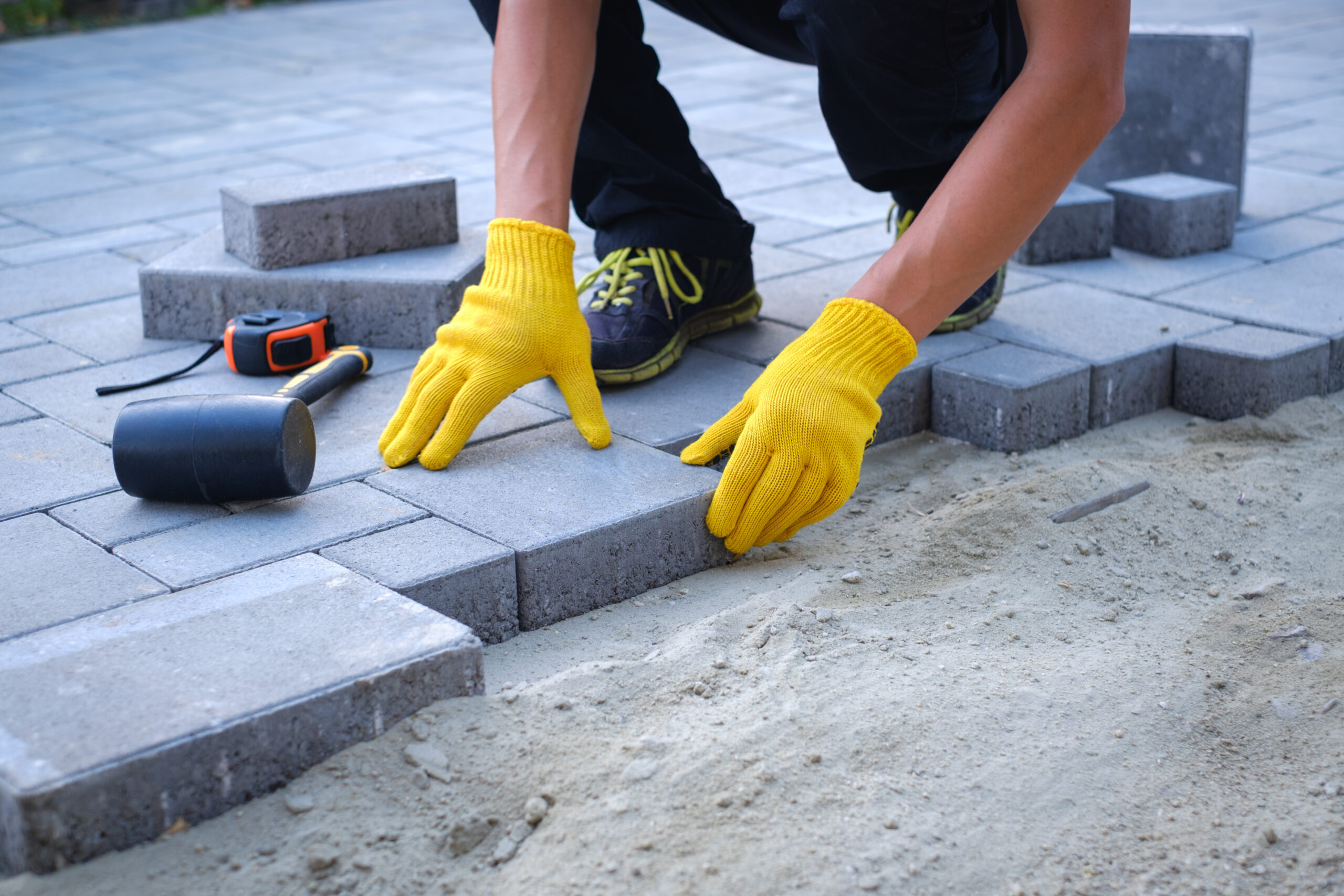 Buttrose Landscaping - person in yellow gloves lays paving stones in layers. Garden brick pathway paving by professional paver worker. Laying gray concrete paving slabs in house courtyard on sand foundation base.